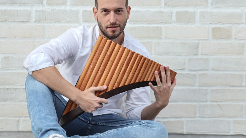 Ein Mann in weißem Hemd und Jeans sitzt vor einer weißen Backsteinwand und hält eine Panflöte in den Händen., © Kevin Schäfer Ein Mann in weißem Hemd und Jeans sitzt vor einer weißen Backsteinwand und hält eine Panflöte in den Händen., © Kevin Schäfer