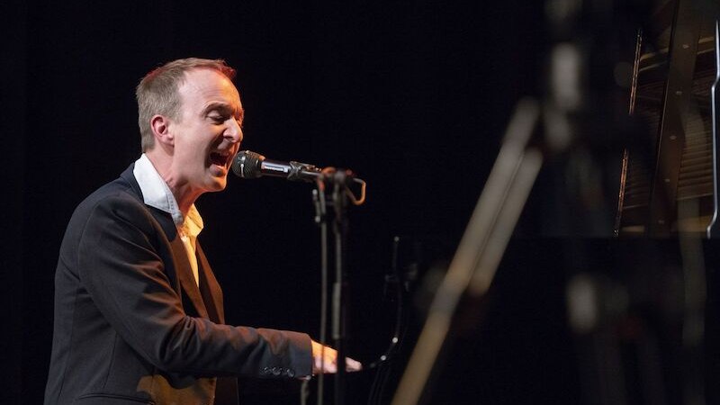 Ein Mann singt leidenschaftlich am Klavier auf einer B&uuml;hne. Er tr&auml;gt einen dunklen Anzug und ist in ein Mikrofon vertieft., &copy; Interessengemeinschaft Kultur Sindelfingen / B&ouml;blingen e.V.