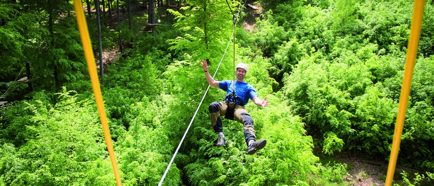 Person mit Helm und Kletterausr&uuml;stung schwebt an einem Seil &uuml;ber einem dichten, gr&uuml;nen Wald im Kletterwald Plochingen., &copy; Sebastian Berger