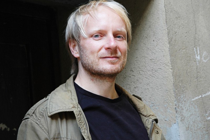 Ein Mann mit blonden Haaren und Bart steht lächelnd vor einer grauen Wand. Er trägt eine olivgrüne Jacke und ein schwarzes T-Shirt., © Kulturamt der Stadt Esslingen am Neckar