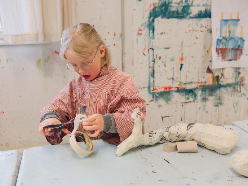Ein Kind in einem gestreiften Kittel bastelt konzentriert mit Klebeband an einem Tisch in einem kreativen Workshop-Raum., © Kunstschule Labyrinth Ein Kind in einem gestreiften Kittel bastelt konzentriert mit Klebeband an einem Tisch in einem kreativen Workshop-Raum., © Kunstschule Labyrinth