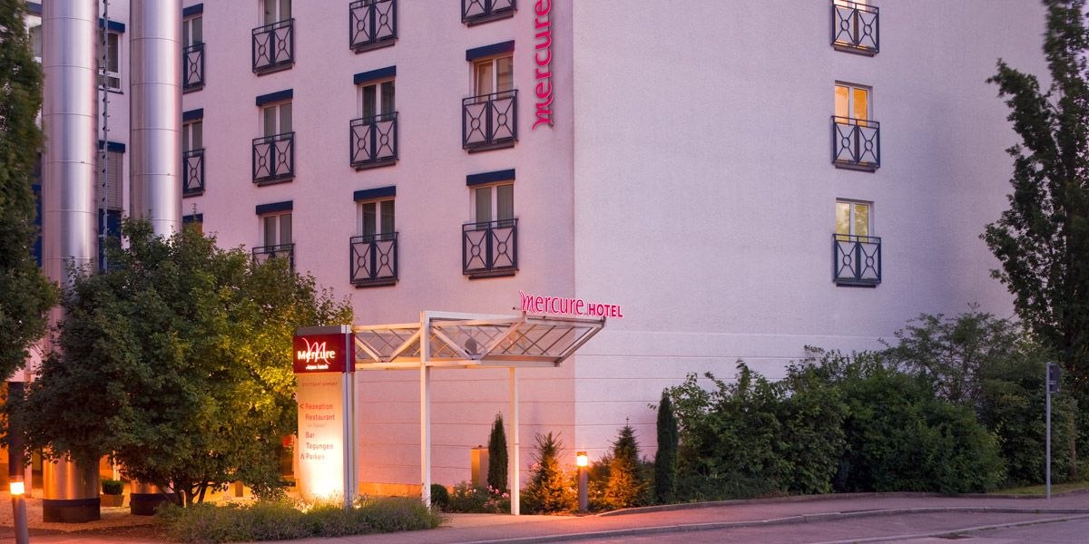 Außenansicht eines Mercure Hotels mit beleuchtetem Eingang, umgeben von Bäumen und Sträuchern., © Mercure Stuttgart Airport Messe Außenansicht eines Mercure Hotels mit beleuchtetem Eingang, umgeben von Bäumen und Sträuchern., © Mercure Stuttgart Airport Messe