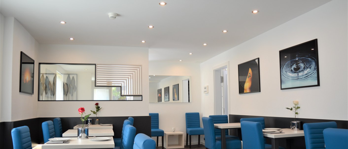 Moderner Frühstücksraum mit blauen Stühlen, weißen Tischen und dekorativen Bildern an den Wänden. Dezente Beleuchtung und stilvolle Dekoration., © Hotel BaWü