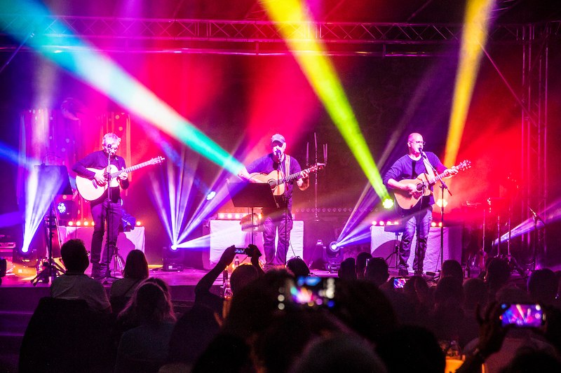 Drei Musiker mit Akustikgitarren auf einer Bühne, umgeben von bunten Lichteffekten. Das Publikum schaut zu und macht Fotos., © Scala Kultur Live gGmbH