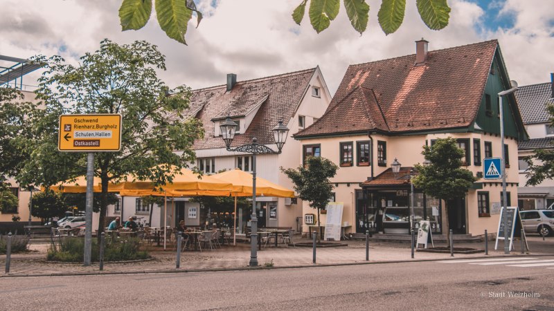 Gemütliche Straßenszene mit Fachwerkhäusern, gelben Sonnenschirmen und einem Café. Ein Wegweiser zeigt Richtungen an. Blätter hängen im Vordergrund., © Stadt Welzheim