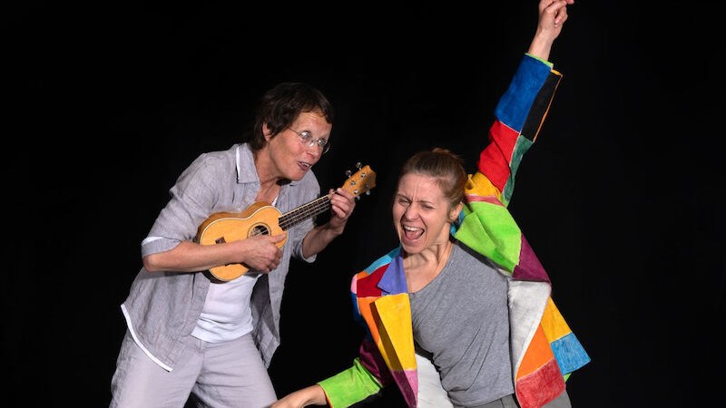 Zwei Personen auf einer Bühne. Eine spielt Ukulele, die andere trägt eine bunte Jacke und tanzt begeistert., © Stadt Winnenden