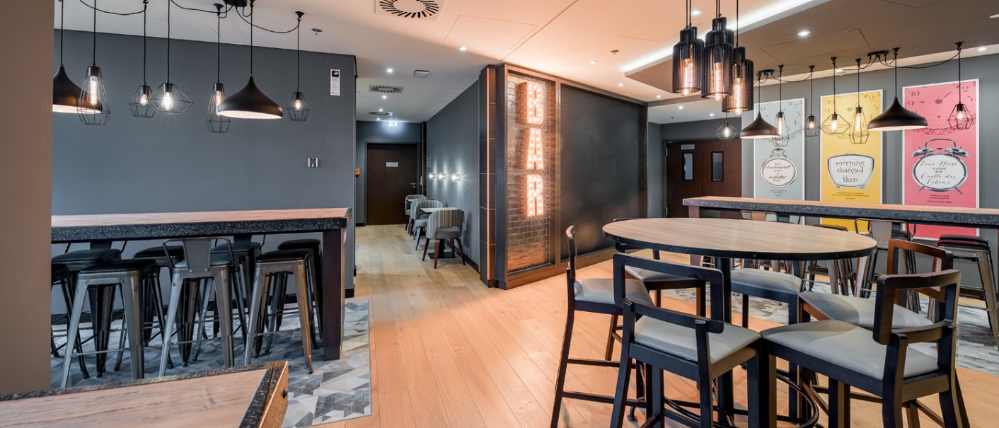 Moderner Frühstücksbereich mit hohen Tischen, Stühlen, stilvollen Lampen und einem leuchtenden 'BAR'-Schild an der Wand., © Premier Inn