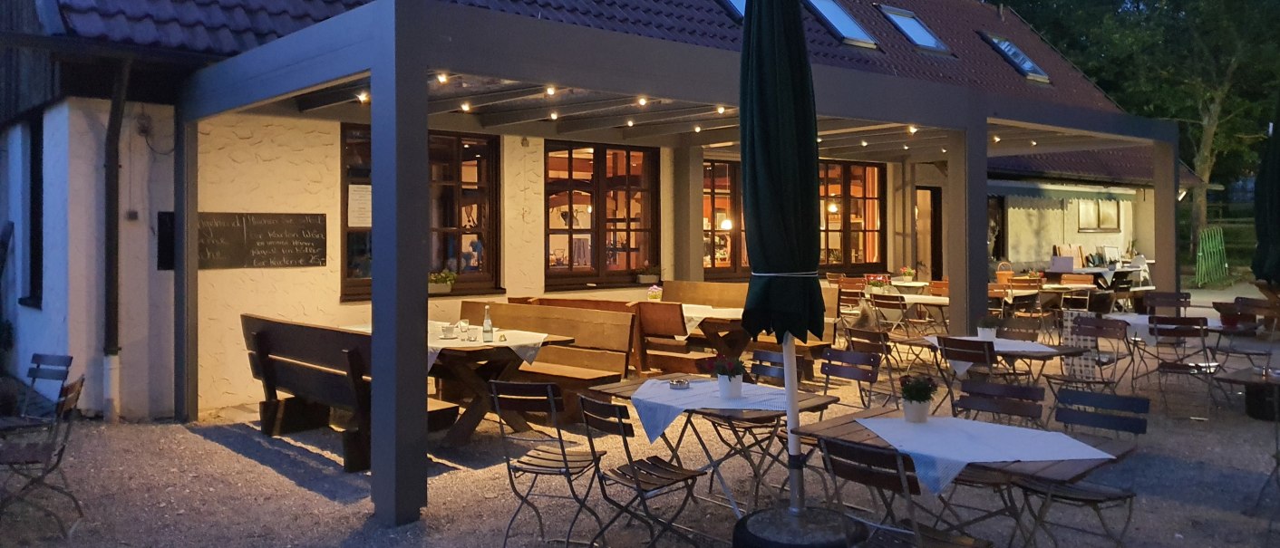 Gemütlicher Außenbereich eines Restaurants bei Dämmerung, mit Holztischen und -stühlen unter einem überdachten Bereich. Beleuchtung sorgt für eine warme Atmosphäre., © Kräuterbühl Gemütlicher Außenbereich eines Restaurants bei Dämmerung, mit Holztischen und -stühlen unter einem überdachten Bereich. Beleuchtung sorgt für eine warme Atmosphäre., © Kräuterbühl