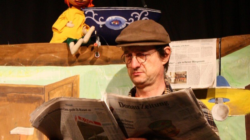 Ein Mann mit Brille und M&uuml;tze liest eine Zeitung auf einer B&uuml;hne. Eine Puppe mit orangefarbenem Haar sitzt auf einem Regal im Hintergrund., &copy; Mauerwerk GmbH