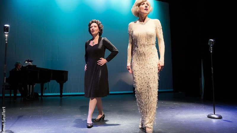 Zwei Frauen stehen auf einer Bühne, eine trägt ein schwarzes Kleid, die andere ein weißes. Im Hintergrund sind Mikrofone und ein Klavier zu sehen., © Stadt Kornwestheim Kulturmanagement Zwei Frauen stehen auf einer Bühne, eine trägt ein schwarzes Kleid, die andere ein weißes. Im Hintergrund sind Mikrofone und ein Klavier zu sehen., © Stadt Kornwestheim Kulturmanagement