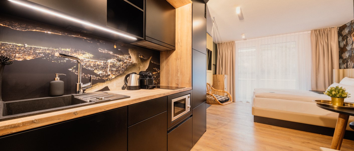 Modernes Apartment mit offener Küche, Holzboden und gemütlichem Schlafbereich. Schwarze Küchenschränke und ein großes Bett dominieren den Raum., © Rioca Stuttgart Posto 4 GmbH