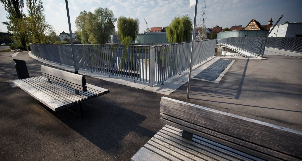 Moderne Brücke mit Metallgeländer, Holzbänken und Bäumen im Hintergrund. Klare, sonnige Atmosphäre mit leicht bewölktem Himmel., © Stoppel Moderne Brücke mit Metallgeländer, Holzbänken und Bäumen im Hintergrund. Klare, sonnige Atmosphäre mit leicht bewölktem Himmel., © Stoppel