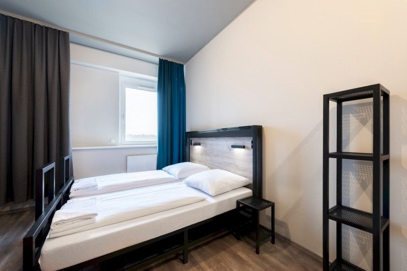 Modernes Doppelzimmer mit zwei Einzelbetten, einem kleinen Nachttisch und einem Regal. Helle Wände und Vorhänge in Blau und Grau., © A&O Hostels Marketing GmbH