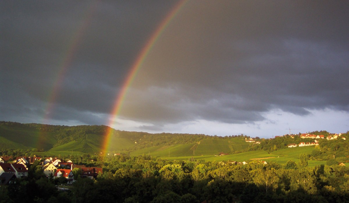 Doppelter Regenbogen über grüner Landschaft mit Häusern, dunklen Wolken und Sonnenlicht auf den Hügeln., © Stuttgart-Marketing GmbH Doppelter Regenbogen über grüner Landschaft mit Häusern, dunklen Wolken und Sonnenlicht auf den Hügeln., © Stuttgart-Marketing GmbH