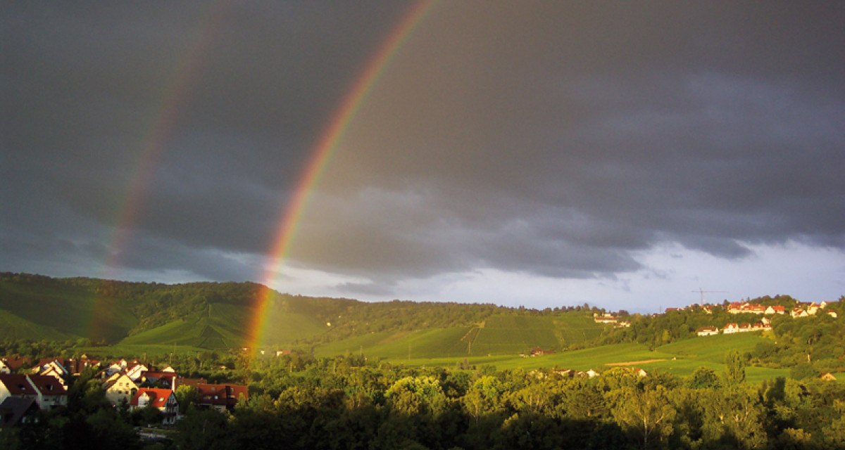Doppelter Regenbogen über grüner Landschaft mit Häusern, dunklen Wolken und Sonnenlicht auf den Hügeln., © Stuttgart-Marketing GmbH Doppelter Regenbogen über grüner Landschaft mit Häusern, dunklen Wolken und Sonnenlicht auf den Hügeln., © Stuttgart-Marketing GmbH