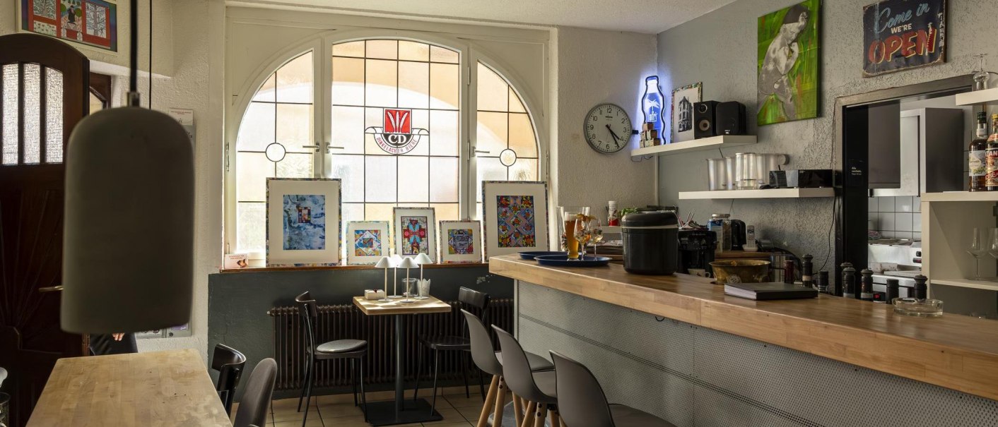 Gemütliches Café mit Holztresen, Kunstwerken an den Wänden und einem großen Fenster. Eine Uhr und Dekorationen schmücken den Raum., © SMG, Sarah Schmid