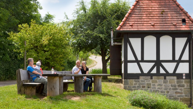 Drei Personen sitzen an einem Holztisch neben einem kleinen Fachwerkhaus im Gr&uuml;nen. Es ist ein sonniger Tag., &copy; Corinna Jacobs
