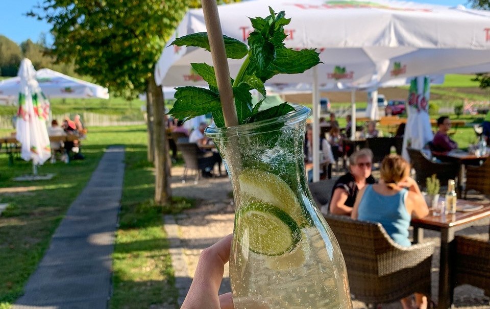 Eine Hand hält eine Karaffe mit Limonade, Minze und Limette. Im Hintergrund sitzen Menschen unter Sonnenschirmen im Freien., © Engel/ Rammerthof