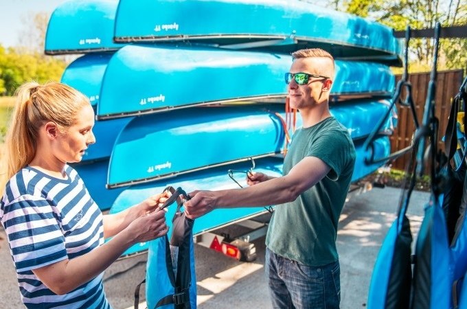 Zwei Personen stehen vor einem Stapel blauer Kanus. Sie halten Schwimmwesten und bereiten sich auf eine Kanutour vor., &copy; Cool-Tours StattReisen