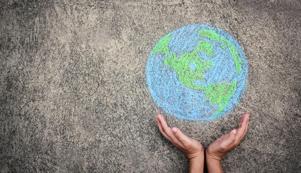 Zwei H&auml;nde strecken sich einer mit Kreide gezeichneten Weltkugel auf Asphalt entgegen, symbolisieren Schutz und Verantwortung f&uuml;r die Erde., &copy; Mit Kreide gemalte Weltkugel auf Asphalt, die von H&auml;nden gerahmt wird.