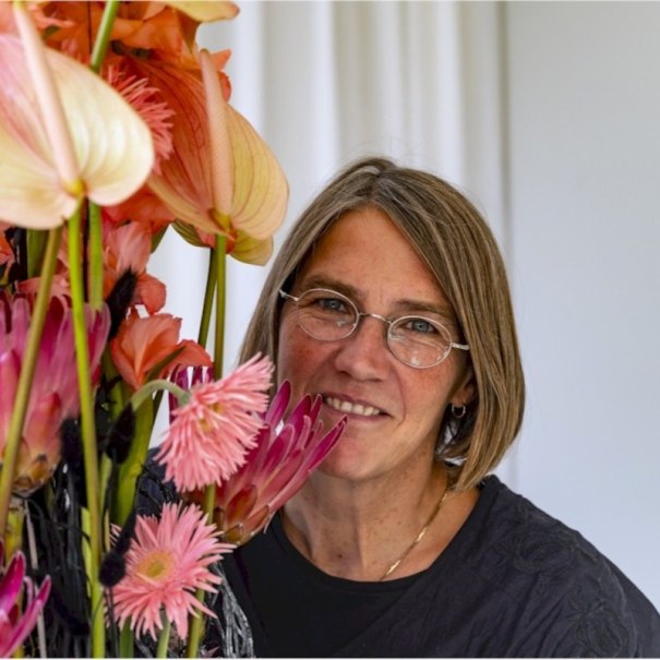 Eine Frau mit Brille l&auml;chelt neben einem bunten Blumenarrangement aus rosa und orangefarbenen Blumen., &copy; Mercedes-Benz AG