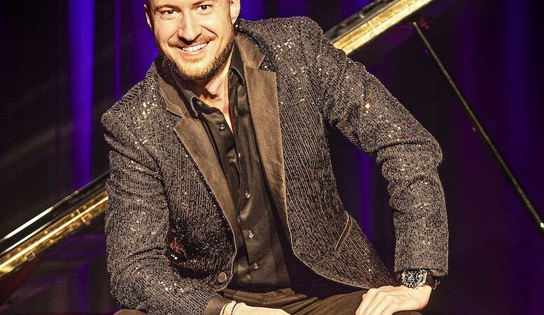 Ein Mann in einem glitzernden Anzug sitzt l&auml;chelnd vor einem Fl&uuml;gel auf einer B&uuml;hne mit violettem Hintergrund., &copy; Stadthalle Leonberg