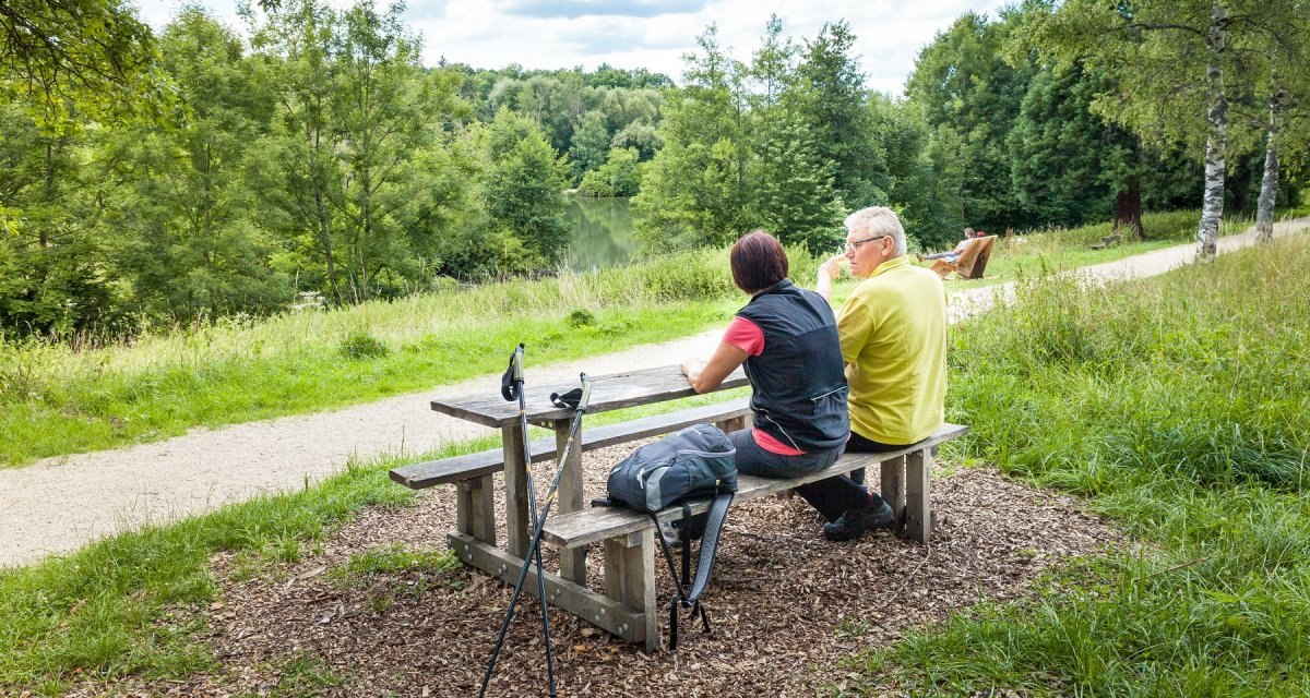 Zwei Personen sitzen an einem Holztisch im Gr&uuml;nen, mit Wanderst&ouml;cken und Rucksack. Sie blicken auf einen See, umgeben von B&auml;umen und einem Weg., &copy; hochgehberge