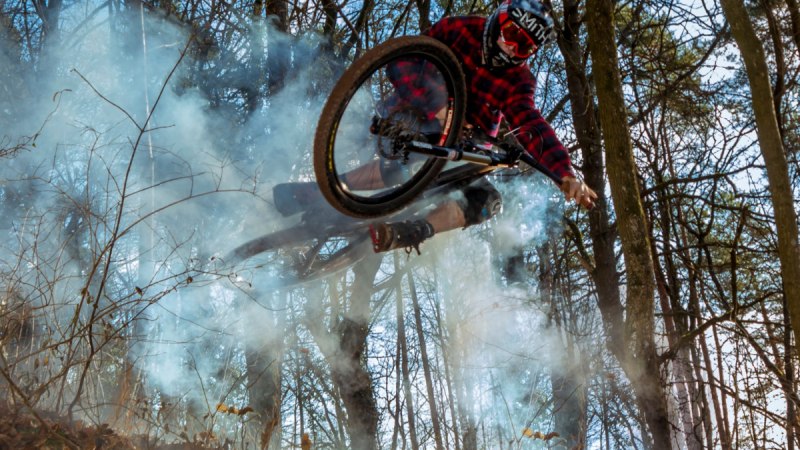 Ein Mountainbiker in rotem Karohemd springt durch den Wald, umgeben von Rauch. Die Szene wirkt dynamisch und abenteuerlich., &copy; Natur.Nah. Sch&ouml;nbuch & Heckeng&auml;u