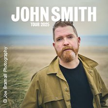 Ein Mann mit Bart steht vor einem Strand. Oben steht 'John Smith Tour 2025'. Fotografiert von Joe Bramall., © links im Bild