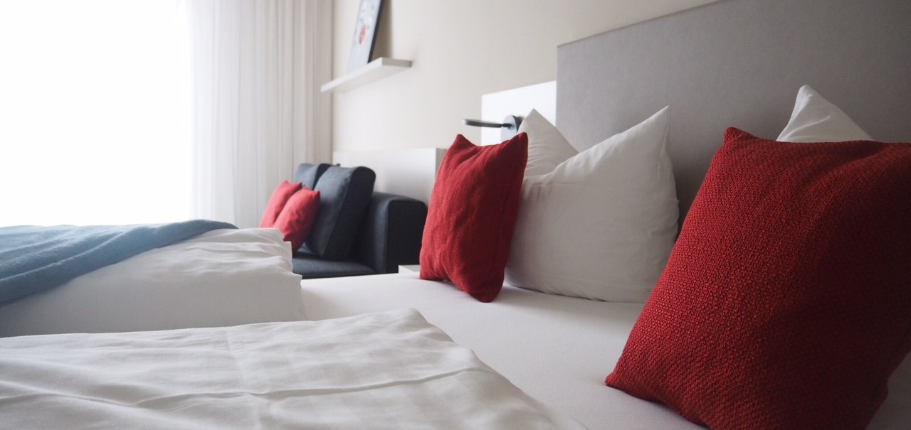 Modernes Hotelzimmer mit weißen Bettlaken, roten Kissen und einem Sofa. Ein Bild hängt an der Wand, helle Vorhänge lassen Licht herein., © TOMAS Modernes Hotelzimmer mit weißen Bettlaken, roten Kissen und einem Sofa. Ein Bild hängt an der Wand, helle Vorhänge lassen Licht herein., © TOMAS