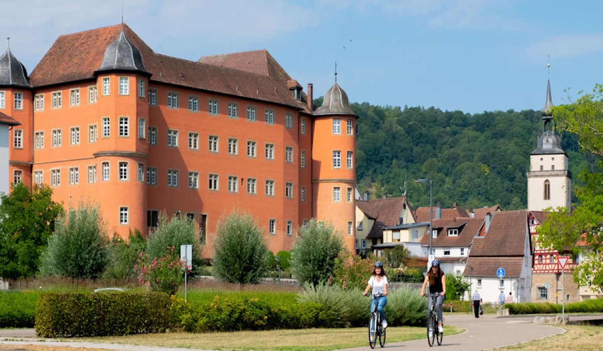 Das Bild zeigt das Schloss in Künzelsau mit seiner markanten roten Fassade. Im Vordergrund fahren zwei Personen auf Fahrrädern. Im Hintergrund ist eine Kirche sichtbar., © Stadt Künzelsau / Olivier Schniepp