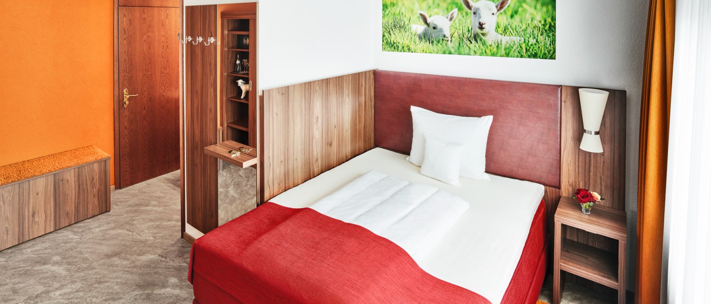 Ein gemütliches Einzelzimmer mit rotem Bett, oranger Wand und einem Bild von Lämmern. Holzmöbel und eine kleine Vase mit Blumen ergänzen das Design., © Hotel Restaurant Lamm