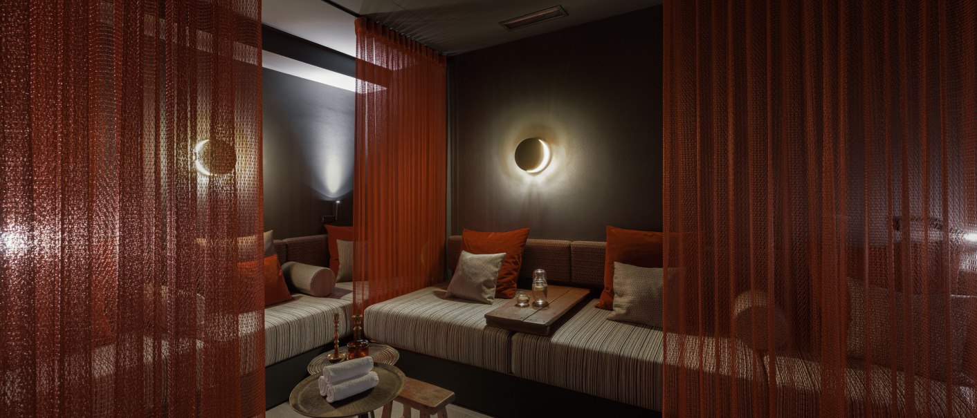 Ein gemütlicher Ruheraum mit roten Vorhängen, gedämpfter Beleuchtung und bequemen Sitzgelegenheiten. Ein Tisch mit Handtüchern und Gläsern steht bereit., © EmiLu Hotel GmbH Ein gemütlicher Ruheraum mit roten Vorhängen, gedämpfter Beleuchtung und bequemen Sitzgelegenheiten. Ein Tisch mit Handtüchern und Gläsern steht bereit., © EmiLu Hotel GmbH