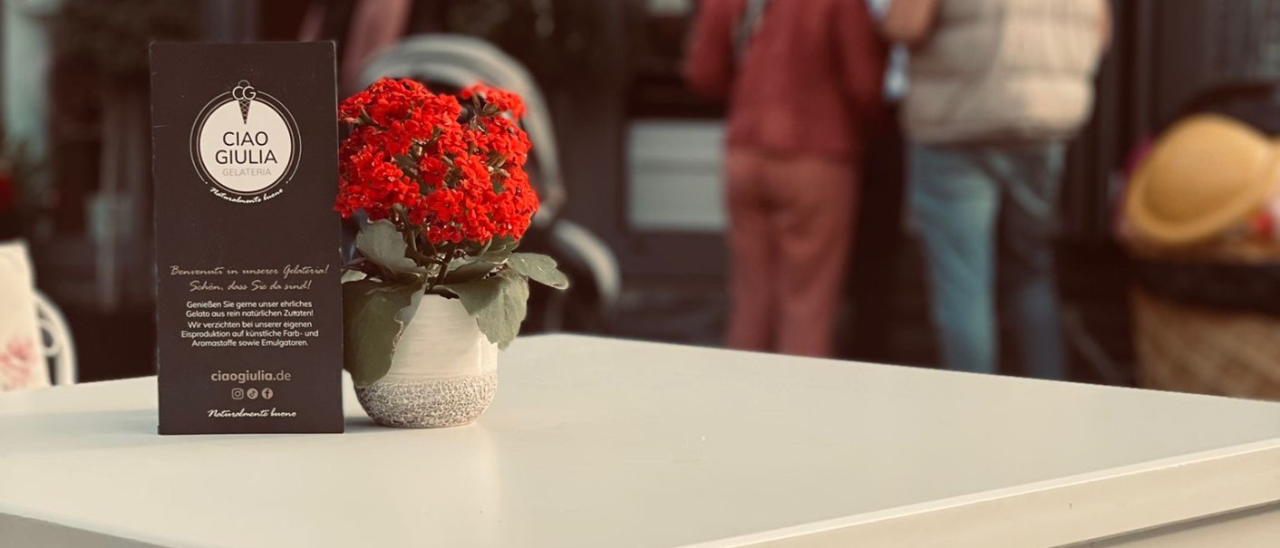 Ein weißer Tisch mit roter Blume steht vor der Eisdiele 'Ciao Giulia'. Im Hintergrund sind Menschen zu sehen., © Ciao Giulia Ein weißer Tisch mit roter Blume steht vor der Eisdiele 'Ciao Giulia'. Im Hintergrund sind Menschen zu sehen., © Ciao Giulia