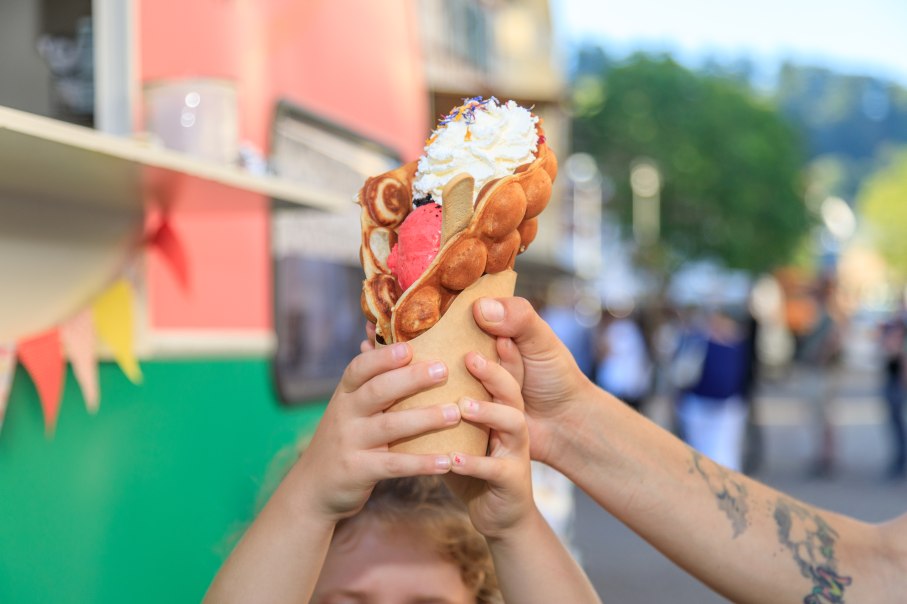 Ein Kind hält eine große Waffel mit Eis und Sahne in den Händen. Im Hintergrund ist ein Foodtruck und eine belebte Straße zu sehen. Ein Kind hält eine große Waffel mit Eis und Sahne in den Händen. Im Hintergrund ist ein Foodtruck und eine belebte Straße zu sehen.