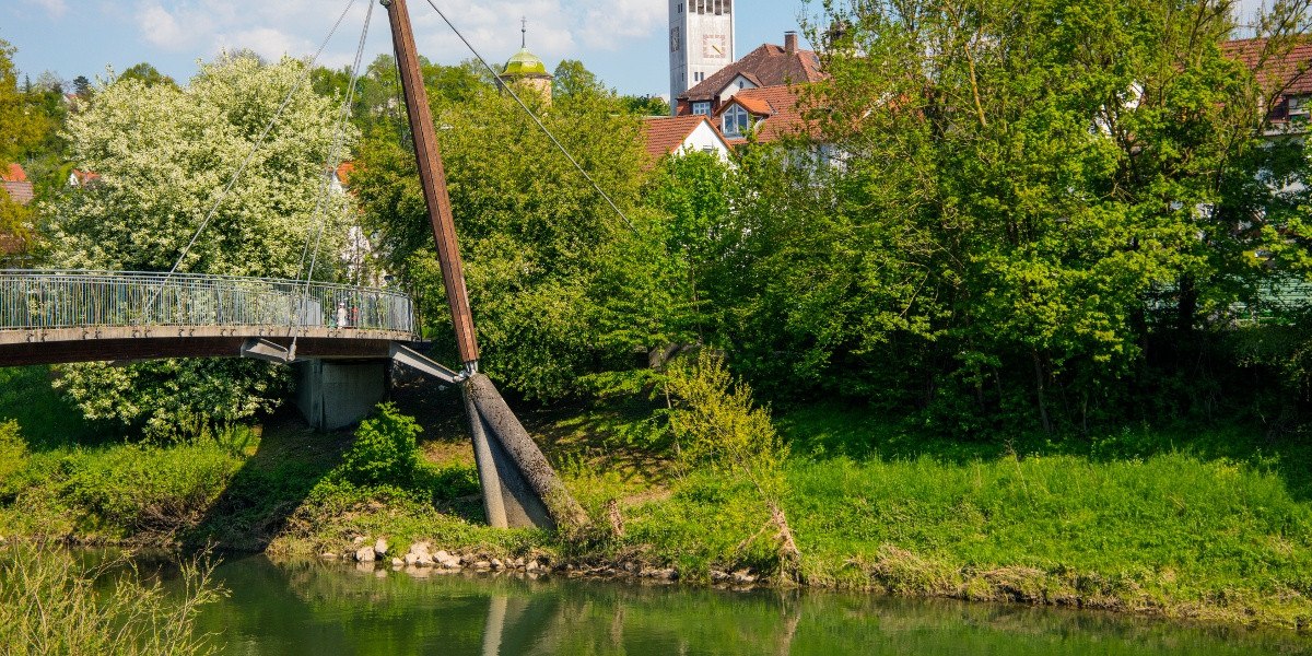 Fußgängerbrücke über einen Fluss, umgeben von grünen Bäumen. Im Hintergrund ist ein Kirchturm zu sehen. Die Szene wirkt idyllisch und ruhig., © Stuttgart-Marketing GmbH Fußgängerbrücke über einen Fluss, umgeben von grünen Bäumen. Im Hintergrund ist ein Kirchturm zu sehen. Die Szene wirkt idyllisch und ruhig., © Stuttgart-Marketing GmbH