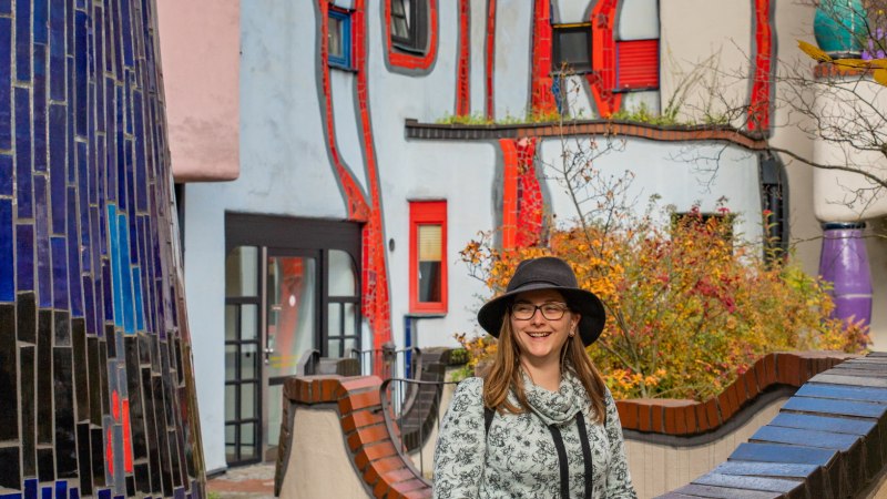 Frau mit Hut vor dem farbenfrohen Hundertwasserhaus in Plochingen, umgeben von herbstlichen Blättern., © Bildergalerie Attila