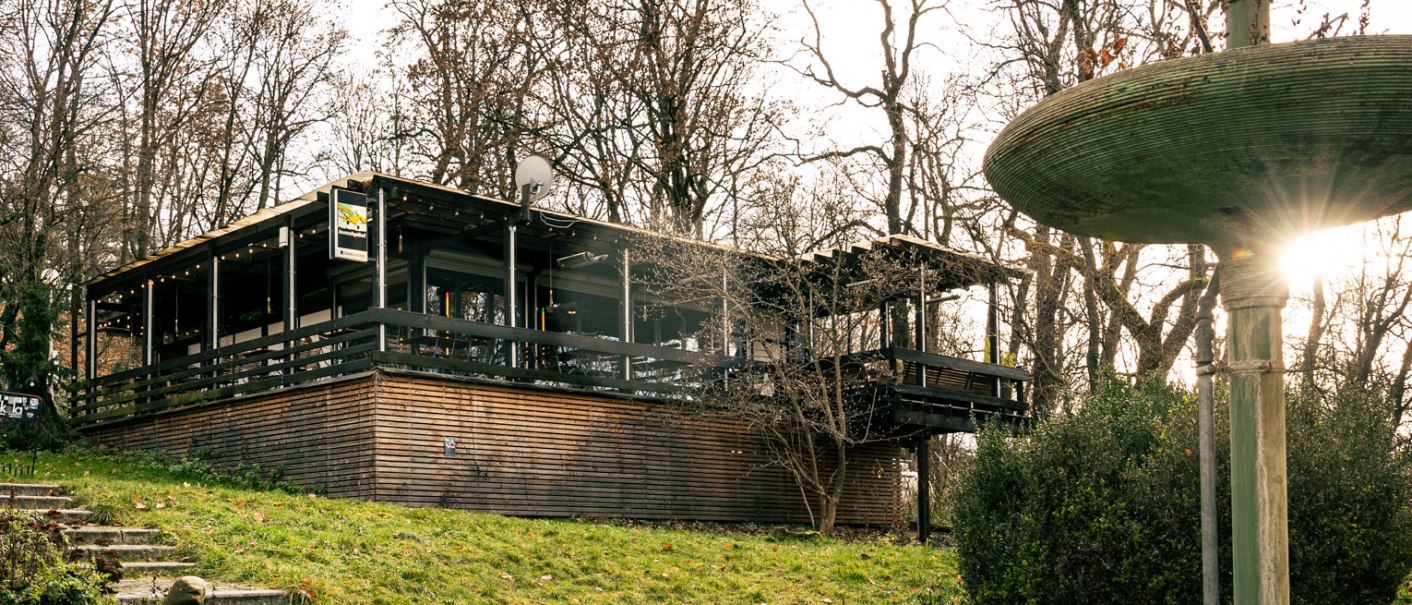 Gebäude mit Holzverkleidung und Terrasse in bewaldeter Umgebung, daneben ein großer, mehrstufiger Brunnen. Sonnenstrahlen scheinen durch die Bäume., © Stuttgart Marketing GmbH, Sarah Schmid Gebäude mit Holzverkleidung und Terrasse in bewaldeter Umgebung, daneben ein großer, mehrstufiger Brunnen. Sonnenstrahlen scheinen durch die Bäume., © Stuttgart Marketing GmbH, Sarah Schmid