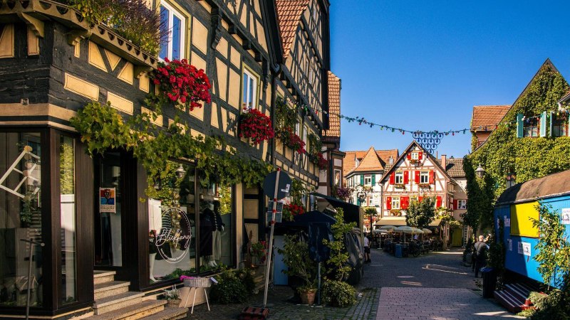 Malerische Altstadtstraße in Besigheim mit Fachwerkhäusern, blühenden Blumen und einem blauen Verkaufswagen unter klarem Himmel., © Stuttgart-Marketing GmbH, Sarah Schmid Malerische Altstadtstraße in Besigheim mit Fachwerkhäusern, blühenden Blumen und einem blauen Verkaufswagen unter klarem Himmel., © Stuttgart-Marketing GmbH, Sarah Schmid