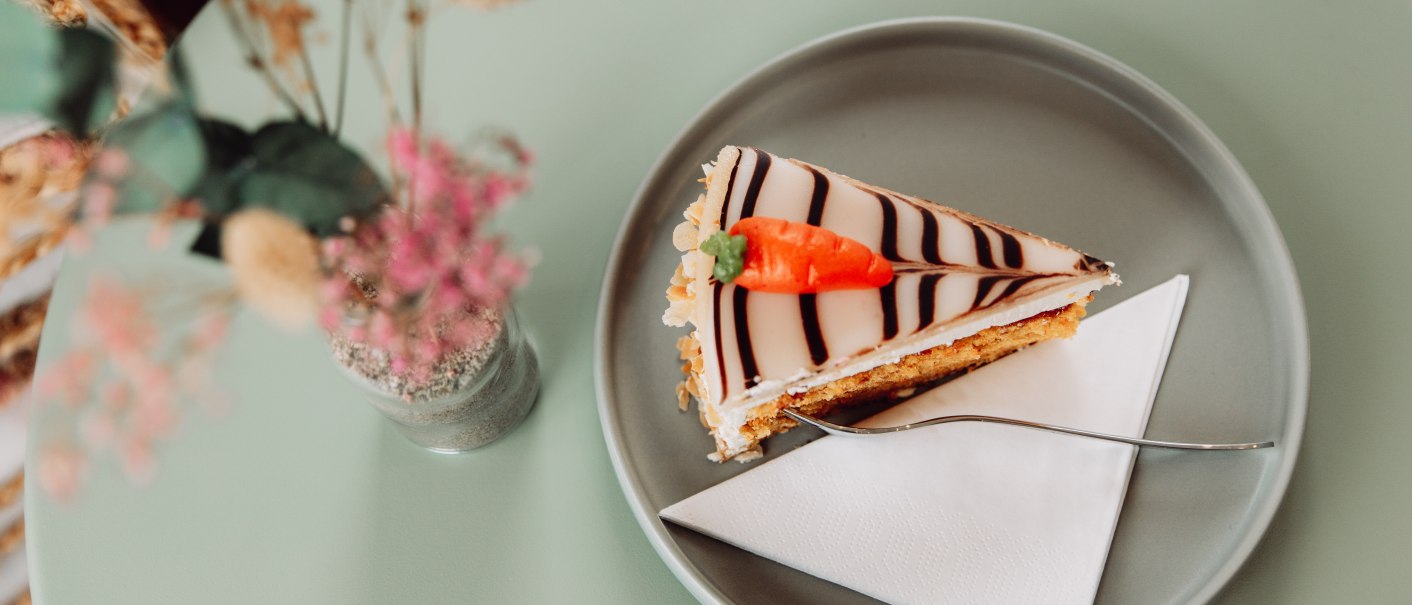 Ein Stück Karottenkuchen mit Zuckerglasur und Marzipankarotte auf einem grauen Teller, daneben eine Vase mit rosa Blumen., © L. Prestle/ Documenting Love Ein Stück Karottenkuchen mit Zuckerglasur und Marzipankarotte auf einem grauen Teller, daneben eine Vase mit rosa Blumen., © L. Prestle/ Documenting Love