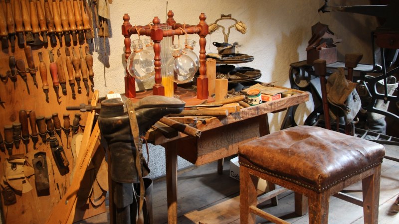 Eine alte Werkstatt im Flößermuseum Calmbach mit Werkzeugen, Lederstiefel und einem Holzhocker. An der Wand hängen zahlreiche Handwerkzeuge., © Touristik Bad Wildbad GmbH