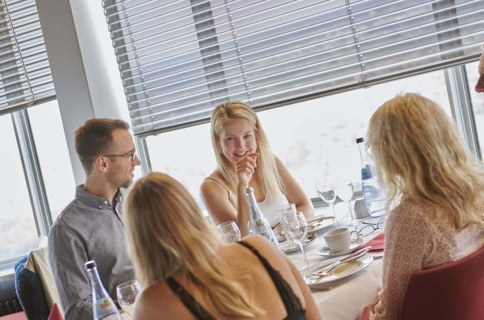 Vier Personen frühstücken an einem Tisch in einem Restaurant mit großen Fenstern. Sie unterhalten sich und genießen die Mahlzeit., © SWR Media Services GmbH / SWR Fernsehturm Stuttgart