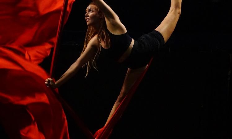 Frau in schwarzer Sportkleidung macht Luftakrobatik mit roten Tüchern in einem dunklen Raum., © Kunstschule Labyrinth Frau in schwarzer Sportkleidung macht Luftakrobatik mit roten Tüchern in einem dunklen Raum., © Kunstschule Labyrinth
