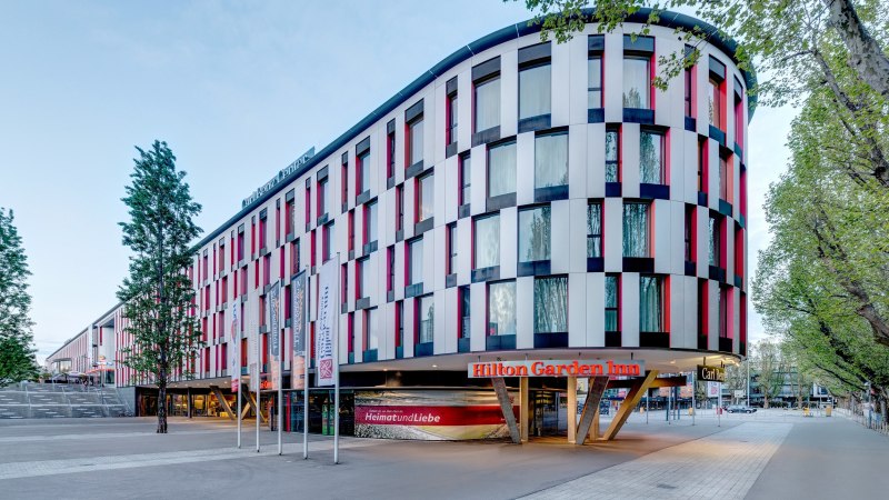 Modernes Hotelgebäude mit bunter Fassade, Hilton Garden Inn Stuttgart Neckarpark. Vor dem Gebäude stehen Fahnenmasten, umgeben von Bäumen., © Hilton Garden Inn Stuttgart Neckarpark Modernes Hotelgebäude mit bunter Fassade, Hilton Garden Inn Stuttgart Neckarpark. Vor dem Gebäude stehen Fahnenmasten, umgeben von Bäumen., © Hilton Garden Inn Stuttgart Neckarpark