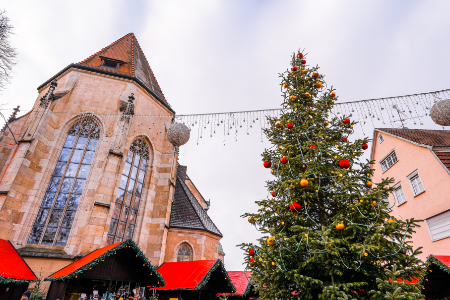 Weihnachtsmarkt mit geschmücktem Baum, Kirche im Hintergrund und Ständen mit roten Dächern. Lichterketten und Kugeln schmücken die Szene., © SMG, L.Martin