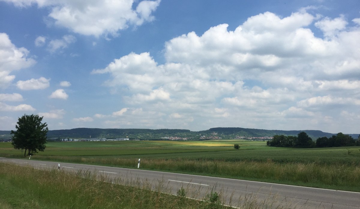 Landschaft mit einer Straße im Vordergrund, Feldern und Hügeln im Hintergrund unter einem blauen Himmel mit Wolken., © www.pro-cycl.de