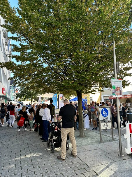 Menschen versammeln sich bei einem Stra&szlig;enfest unter einem gro&szlig;en Baum. Im Hintergrund sind St&auml;nde und ein Schild f&uuml;r eine Fu&szlig;g&auml;ngerzone zu sehen.
