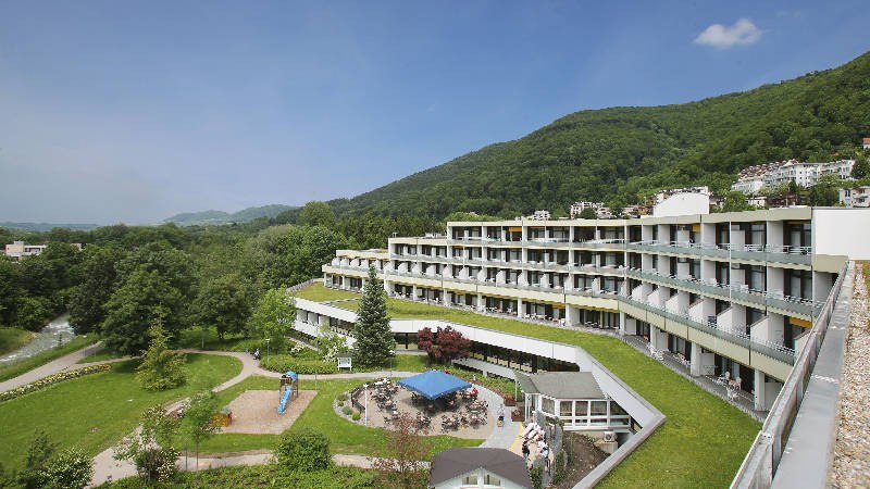 Modernes Gebäude mit Terrassen in grüner Umgebung und Hügeln im Hintergrund. Ein Spielplatz und Sitzbereich sind im Vordergrund sichtbar. Modernes Gebäude mit Terrassen in grüner Umgebung und Hügeln im Hintergrund. Ein Spielplatz und Sitzbereich sind im Vordergrund sichtbar.