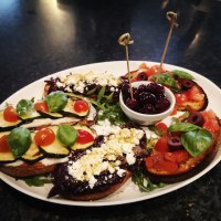 Ein Teller mit Antipasti: Bruschetta mit Tomaten, Zucchini, Oliven und Basilikum, dazu eine Schale mit Oliven in der Mitte., © La buona Forketta