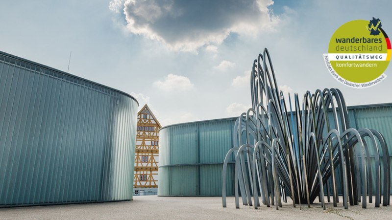 Moderne Skulptur vor einem Fachwerkhaus in Waiblingen. Im Hintergrund Wolken am Himmel. Logo von 'Wanderbares Deutschland' oben rechts., © Unbekannt